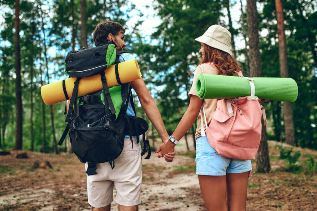 couple with backpacks and sleeping pads hold hands while walking in the woods