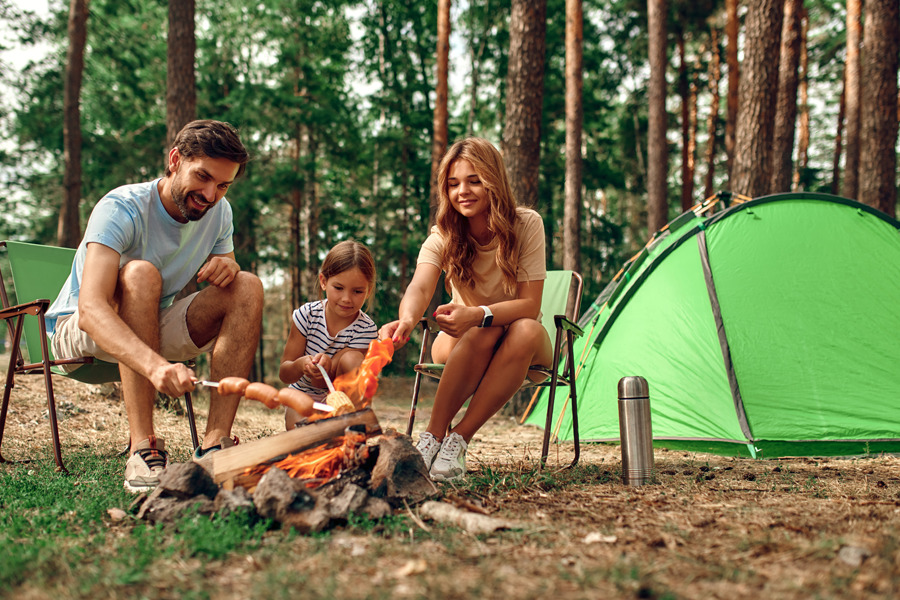 Happy family sitting by the fire near the tent in a pine forest.