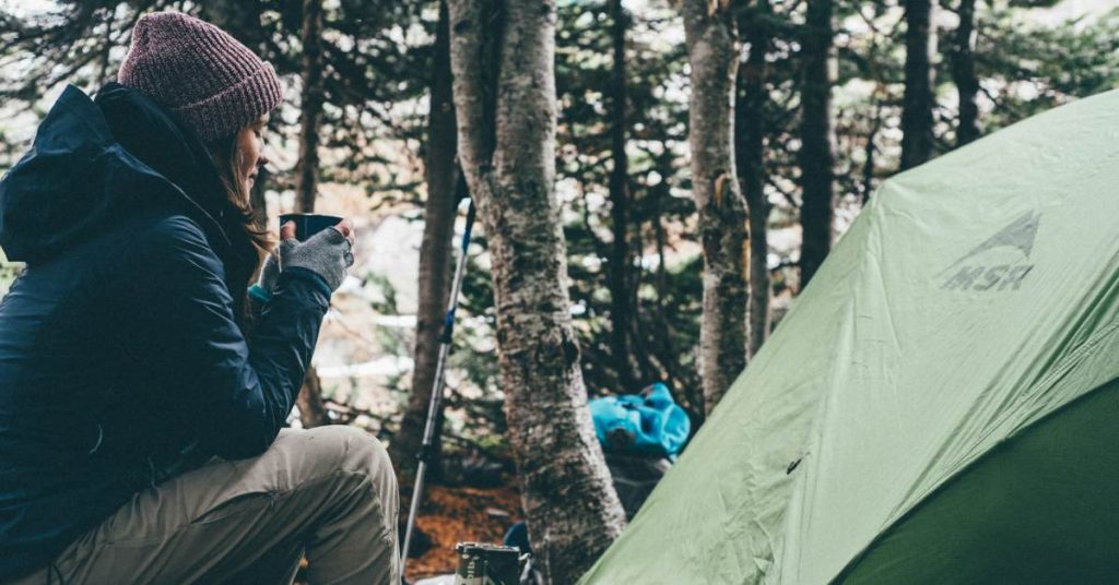 woman camping sitting outside her green tent in a forest wearing warm clothing and holding a mug of coffee