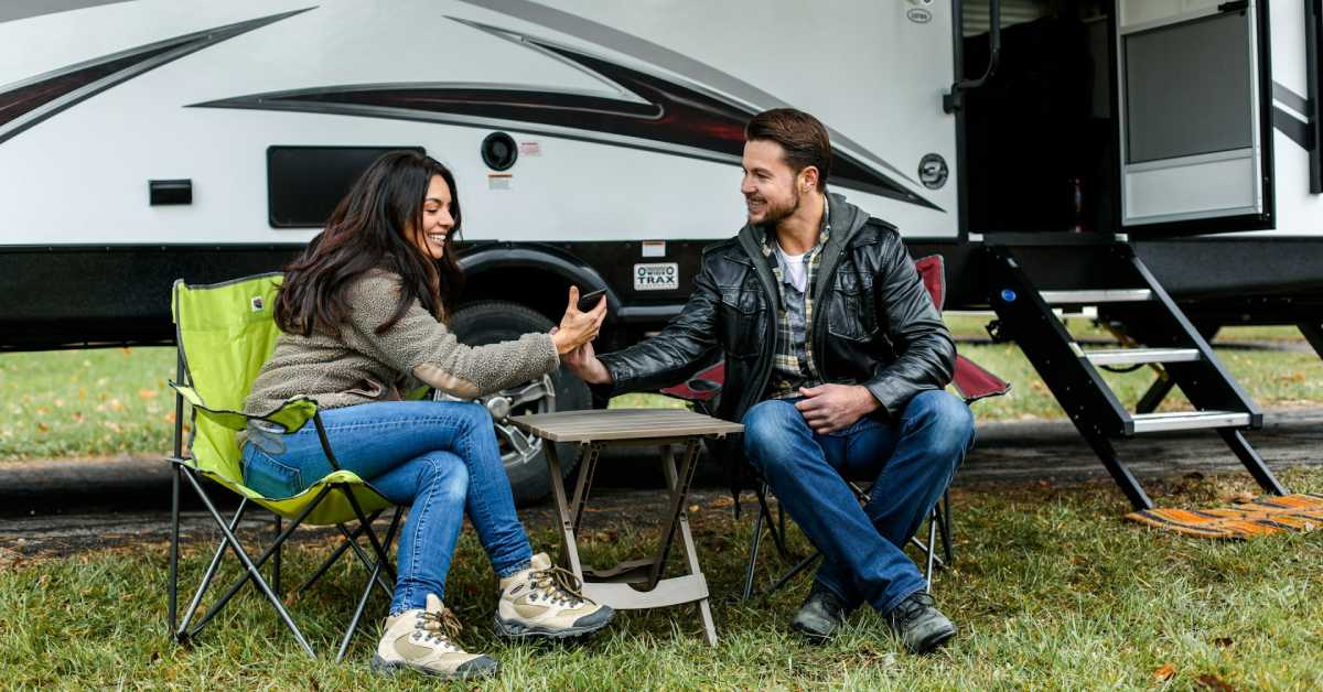 man and woman sitting outside of an rv in bag chairs looking at a phone